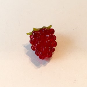 May include: A detailed, red glass raspberry with a cluster of small, round beads. The raspberry has a green stem and leaves. The image is a close-up shot of the raspberry against a white background.