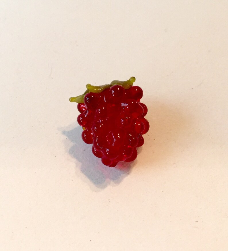 Pode incluir: Uma framboesa de vidro vermelho detalhada com um aglomerado de pequenas contas redondas. A framboesa tem um caule e folhas verdes. A imagem &eacute; um close-up da framboesa contra um fundo branco.