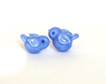 Può includere: Due piccole statuine di uccelli in vetro blu. Gli uccelli si guardano, con i loro becchi quasi a toccarsi.