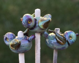 Può includere: Tre statuette di uccelli in vetro nelle tonalità iridescenti del blu, viola e verde, ciascuna con piccoli occhi gialli e ali marrone scuro. Gli uccelli sono montati su singole aste verticali grigio chiaro e presentano una finitura liscia.