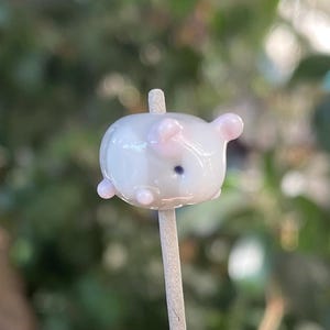 May include: A small, white glass bead shaped like a pig with pink ears and a black nose. The bead is on a wooden stick.