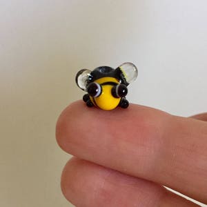 May include: A tiny, handcrafted glass bee figurine. The bee features a yellow body with black stripes, black eyes, and transparent wings. The miniature bee is a charming decorative item.