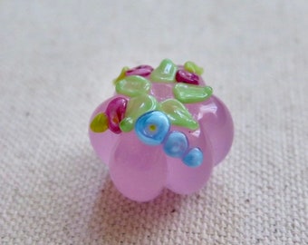 Può includere: Una perla di vetro rosa chiaro fatta a mano con un design floreale. La perla presenta piccoli fiori di vetro colorati nei toni del blu, rosa e verde. La perla è rotonda con una superficie leggermente strutturata, ideale per la creazione di gioielli.