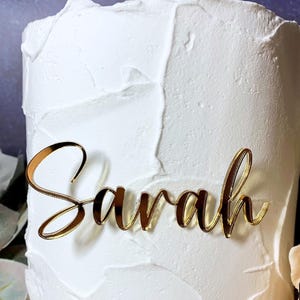Może przedstawiać: Złoty akrylowy topper na tort z imieniem Sarah napisanym kursywą, umieszczony na froncie białego tortu z teksturowanym wykończeniem. Topper ma odblaskową powierzchnię i jest wyśrodkowany na boku tortu.
