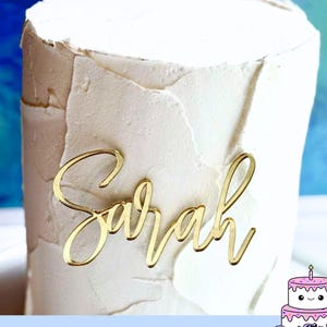May include: A gold-colored acrylic cake topper featuring the name Sarah in a cursive script font, positioned on the front of a white cake with a textured, layered frosting finish. The topper has a reflective, metallic surface.