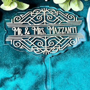 May include: A gold and clear acrylic cake topper with an art deco design. The topper has the text 'Mr. & Mrs. Mazzanti'.