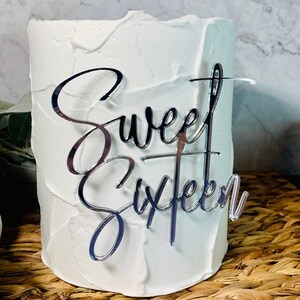 May include: A cylindrical white cake with textured frosting features a reflective silver acrylic cake topper. The topper is written in a cursive script font and displays the text Sweet Sixteen, with a vertical line accenting the letter t.