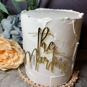 May include: White cake with gold glitter frosting and a gold acrylic topper that says "The Martins".