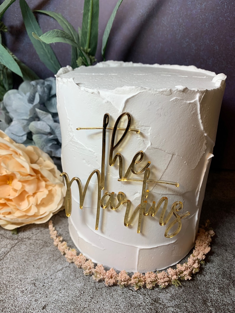 May include: White cake with gold glitter frosting and a gold acrylic topper that says "The Martins".