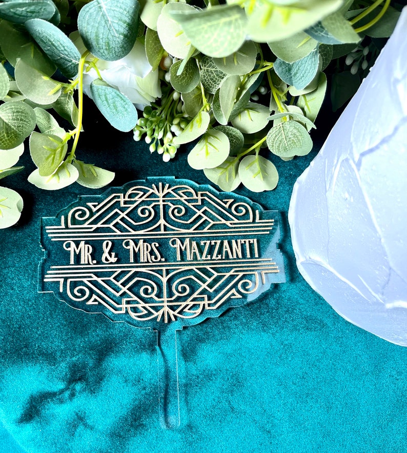 May include: A gold and clear acrylic cake topper with an art deco design. The topper has the text 'Mr. & Mrs. Mazzanti'.