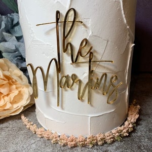 May include: White cake with gold glitter lettering that says "The Martins" on top.