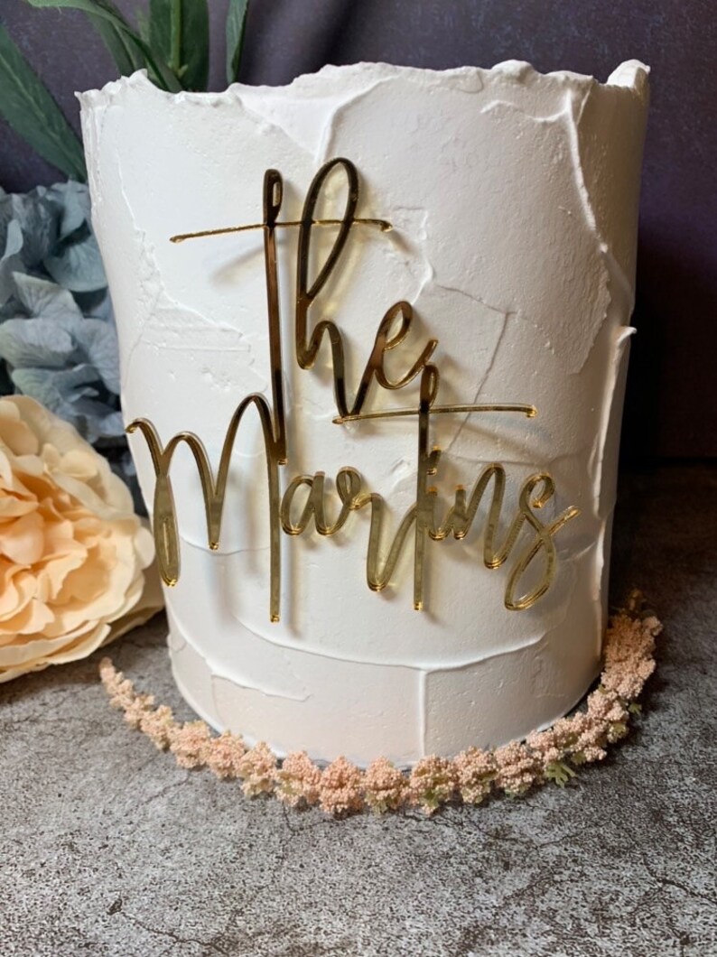 May include: White cake with gold glitter lettering that says "The Martins" on top.
