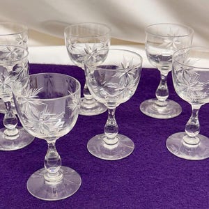 Puede incluir: Conjunto de siete copas de vino de cristal transparente con diseños de estrellas. Cada copa tiene una base redonda, un tallo curvo y una copa grabada con un patrón de estrella. Las copas están dispuestas sobre una superficie púrpura.