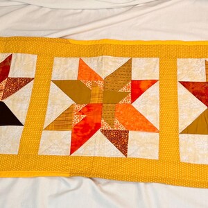 Peut inclure: Une courtepointe rectangulaire faite à la main avec trois motifs d'étoiles. Chaque étoile est un patchwork de tissus orange, marron et crème. La courtepointe a une bordure jaune à motifs de points et est exposée sur une surface blanche.