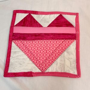 Può includere: Un sottobicchiere quadrato trapuntato con un motivo a cuore. Il sottobicchiere presenta sfumature di rosa e bianco, con un bordo a fantasia. Il cuore è composto da triangoli e strisce orizzontali.