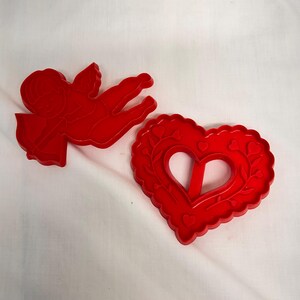May include: Two red plastic cookie cutters. One cutter is shaped like a winged cherub holding a bow and arrow. The second cutter is shaped like a heart with a scalloped edge and an embossed vine pattern featuring small hearts along the border.
