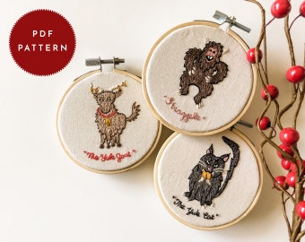 Könnte beinhalten: Drei bestickte Bilder in Ringen mit einem braunen, pelzigen Monster, einer braunen Ziege mit Geweih und einer schwarzen Katze. Das Monster ist mit "The Straggler" beschriftet, die Ziege mit "The Yule Goat" und die Katze mit "The Yule Cat".