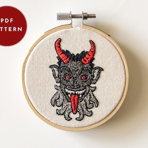 May include: Embroidered hoop art featuring a black and gray devil with red horns and a red tongue. The devil is stitched on a beige fabric and framed in a wooden hoop.