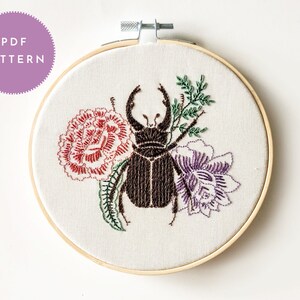 Peut inclure: Motif de broderie PDF dans un cercle en bois. Le motif représente un scarabée brun foncé avec de grandes pinces, entouré de broderies florales rouges, violettes et vertes. Le texte "PDF PATTERN" est dans un cercle violet.