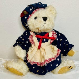 May include: A vintage teddy bear with cream-colored fur, wearing a navy blue dress with white stars and a matching bonnet. The dress has a cream-colored apron with a red bow and trim. The bear is sitting.