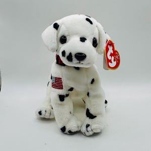 May include: A plush Dalmatian dog toy with black spots on a white body. It has a red collar and a small American flag patch. A red and white tag is attached to the ear.