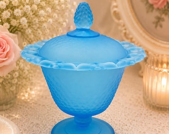 May include: A bright blue frosted glass candy dish with a lid. The bowl features a textured diamond pattern on the exterior, a scalloped rim, and a pedestal base. The matching lid is topped with a small, textured finial shaped like a pinecone.