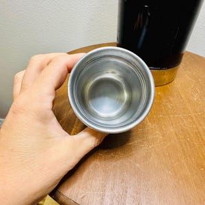 May include: A small, stainless steel cup is held in a hand, with a black container in the background. The cup has a simple, metallic design. The background includes a wooden table and a white wall.