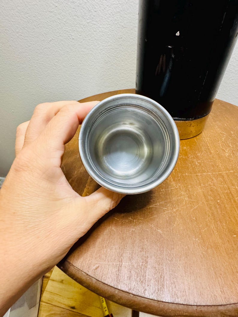 May include: A small, stainless steel cup is held in a hand, with a black container in the background. The cup has a simple, metallic design. The background includes a wooden table and a white wall.