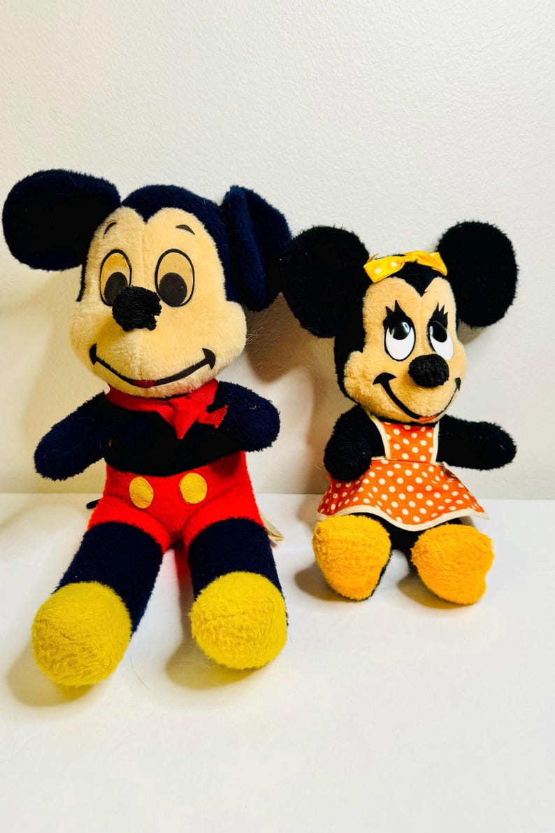 May include: Two plush Disney character dolls, Mickey Mouse and Minnie Mouse. Mickey wears red pants, yellow shoes, and a red bow tie. Minnie wears a polka-dot dress and a yellow bow. Both have black ears.