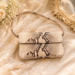 May include: A rectangular shoulder bag featuring a beige and dark brown snakeskin pattern. The bag has a structured flap closure and a matching thin shoulder strap. The exterior surface displays a detailed, textured reptilian scale print.