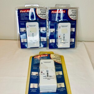 May include: Three all-in-one travel adapters in clear plastic packaging. Each adapter is white with black and red text, featuring universal plugs and USB charging capabilities. The packaging includes the "First Alert" logo and details about worldwide use.