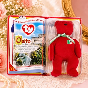 May include: A red plush bear toy named Osito, packaged in a clear plastic display case alongside a booklet titled International Bears II. The bear features a green, white, and red ribbon around its neck and a small Mexican flag patch on its chest.