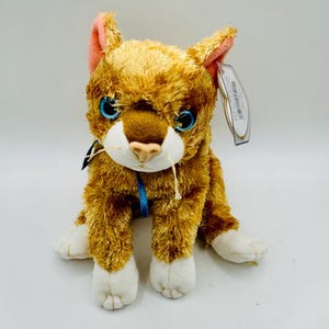 May include: A plush toy cat with orange and brown fur, blue eyes, and pink inner ears. The cat has white paws and a blue ribbon around its neck. A tag is attached to the ear. The cat is sitting upright.