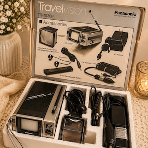 May include: A vintage Panasonic Travelvision TR-1030P portable television set in its original styrofoam packaging. The set includes a black and silver television with an antenna, a detachable hood screen, an earphone, a car adaptor cord, and an AC adaptor. The box lid features product illustrations and text.