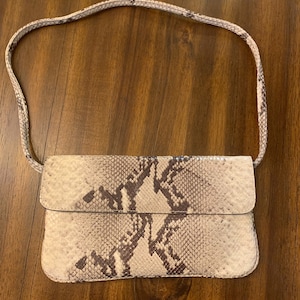 May include: A rectangular handbag with a flap closure, featuring a snakeskin pattern in shades of beige, brown, and gray. The bag has a matching strap. The bag is lying on a wooden surface.