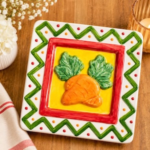 May include: A square ceramic plate featuring a raised orange carrot with green leafy tops centered on a yellow background. The plate is framed by a thick red border and an outer white edge decorated with a green zigzag pattern and red dots.