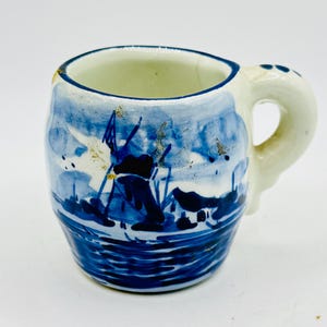 May include: A small ceramic mug with a white base and a hand-painted blue design featuring a windmill landscape. The mug has a thick, rounded handle and a blue rim. The glaze shows minor signs of wear with small gold-colored flecks on the surface.