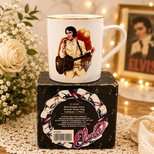 May include: A white ceramic mug with a gold rim featuring a graphic of Elvis Presley in a yellow jumpsuit playing an acoustic guitar. The mug sits on a black box with musical notes and text that reads Elvis Presley Army Portrait Photo Mug.