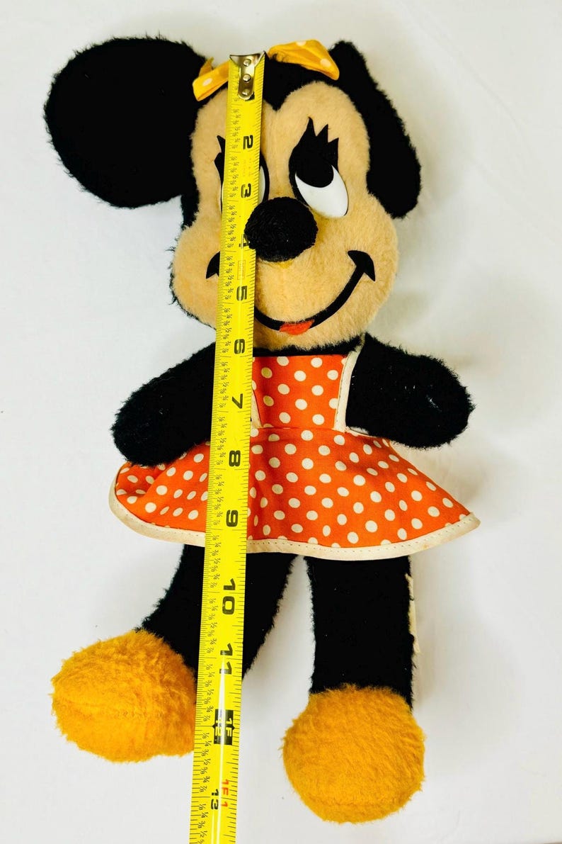 May include: Vintage Minnie Mouse plush doll, approximately 12 inches tall. The doll features black fur, a tan face, and an orange dress with white polka dots. A yellow bow adorns her head. The doll's feet are yellow.
