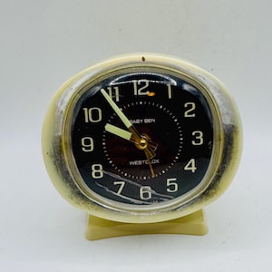 May include: A vintage Baby Ben Westclox alarm clock. The clock has a cream-colored, oval-shaped case with a black face. The face features white numerals and hands, with the words "Baby Ben" and "Westclox" printed on it.