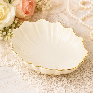 May include: A cream-colored ceramic dish shaped like a shell with a scalloped edge and a thin gold-painted rim. The bowl features a smooth, glossy finish and a shallow, curved interior, providing a decorative vessel for small items or trinkets.