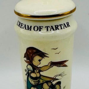 May include: Cream-colored ceramic jar with lid, gold trim, and the words "CREAM OF TARTAR". The jar features a painted illustration of a girl holding a tray, with a small dog and birds. The lid has a gold knob.