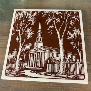 May include: A brown and white ceramic tile with a vintage illustration of a church with two trees on either side. The church has a steeple and a white picket fence in front of it.