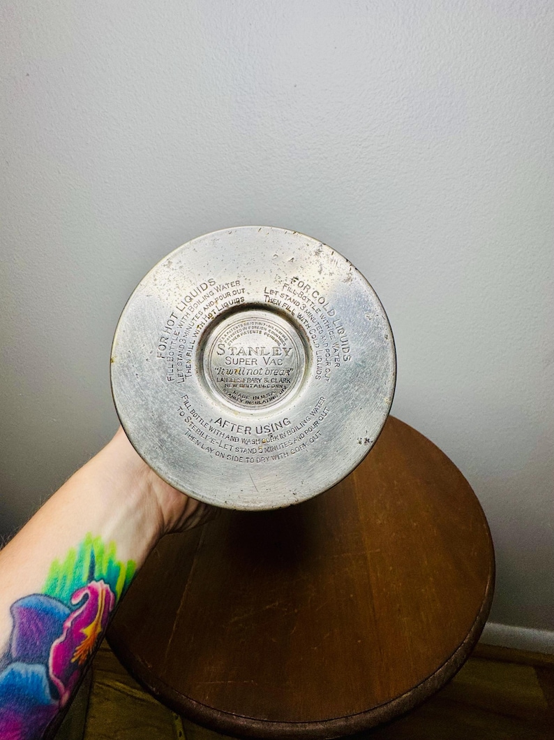 May include: A vintage Stanley Super Vac thermos bottom, featuring instructions for hot and cold liquids. The metal base has circular text and a central logo. The thermos is resting on a wooden surface.