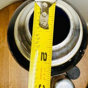 May include: A black and silver container with a blue interior, measured with a yellow ruler. The ruler shows a measurement of approximately 2 inches. The container has a silver rim and a cylindrical shape.