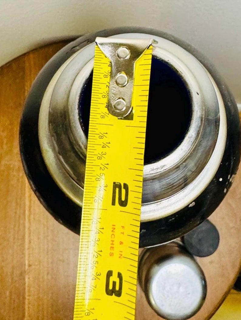 May include: A black and silver container with a blue interior, measured with a yellow ruler. The ruler shows a measurement of approximately 2 inches. The container has a silver rim and a cylindrical shape.