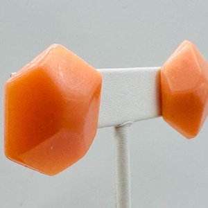 May include: A pair of orange geometric earrings. Each earring has a faceted, angular design. The earrings are displayed on a white earring stand against a light gray background. The earrings have a glossy finish.