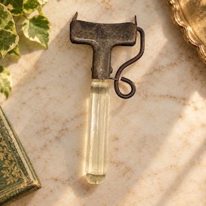 May include: A vintage kitchen peeler featuring a weathered, dark metal T-shaped head and a clear, faceted glass handle. The tool includes a curved metal side arm attached to the main body, showcasing a utilitarian design with visible signs of age.