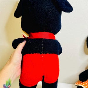 May include: A vintage plush toy of a cartoon character, featuring a black head and ears, red shorts, and a red ribbon. The toy is held by a person. The toy has a black tail.