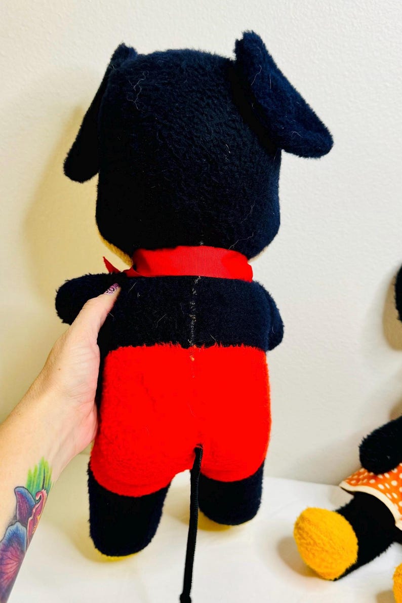 May include: A vintage plush toy of a cartoon character, featuring a black head and ears, red shorts, and a red ribbon. The toy is held by a person. The toy has a black tail.